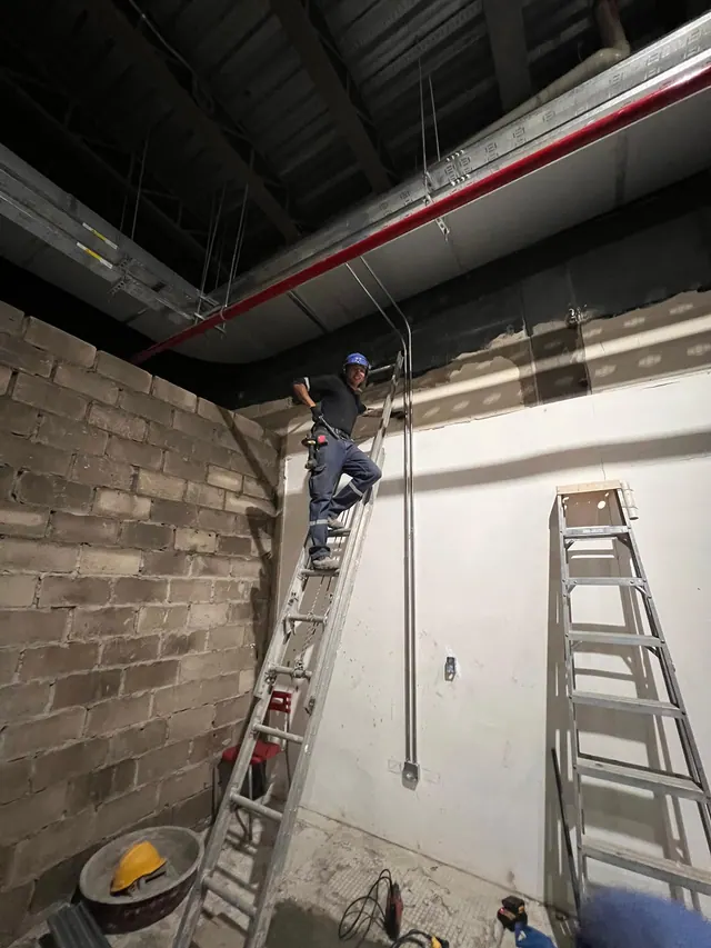 Técnico sobre una escalera junto a canalizaciones y bandejas eléctricas bajo una cubierta metálica